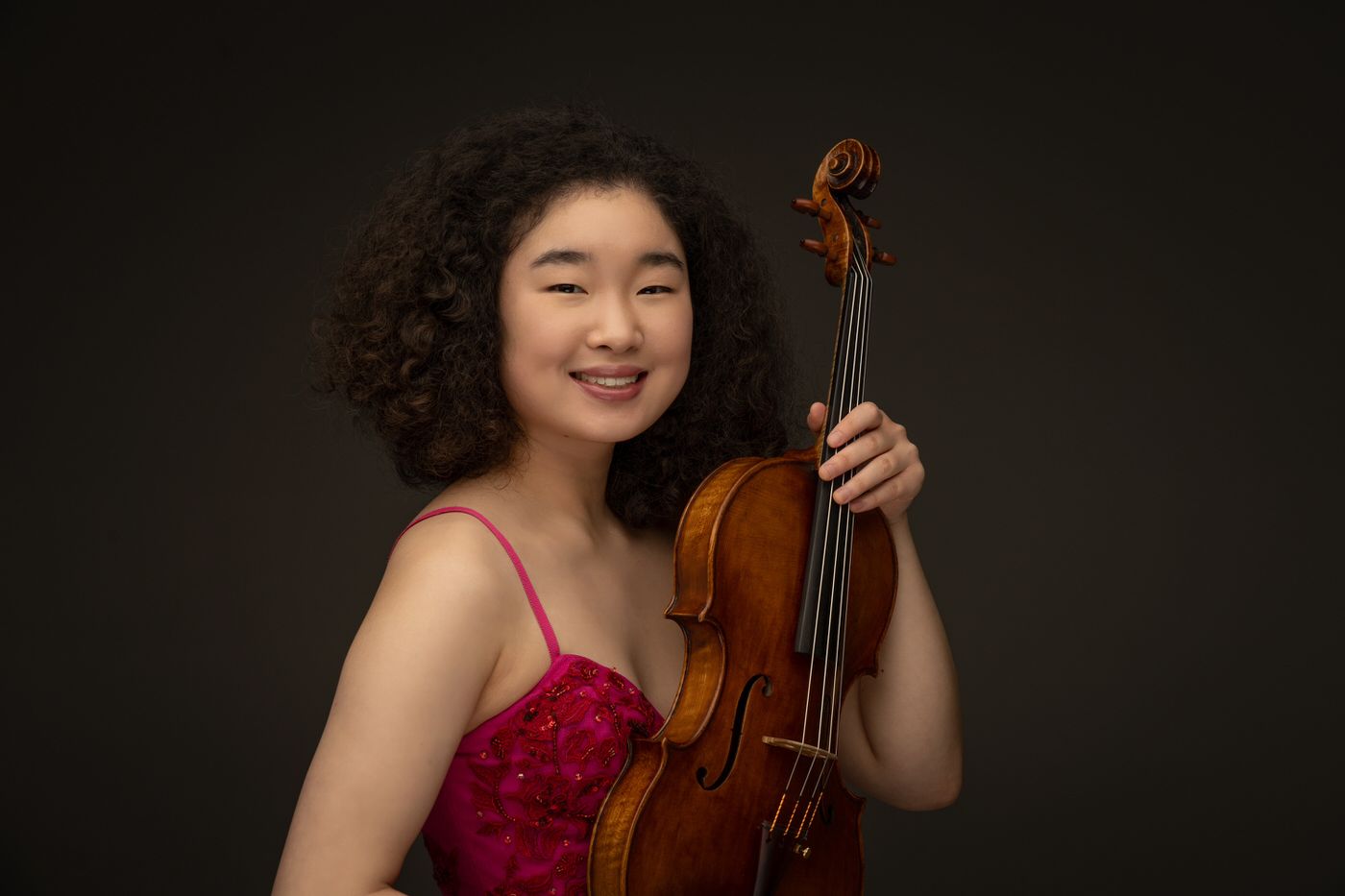 A musician in a red dress holds a violin and smiles against a dark background.