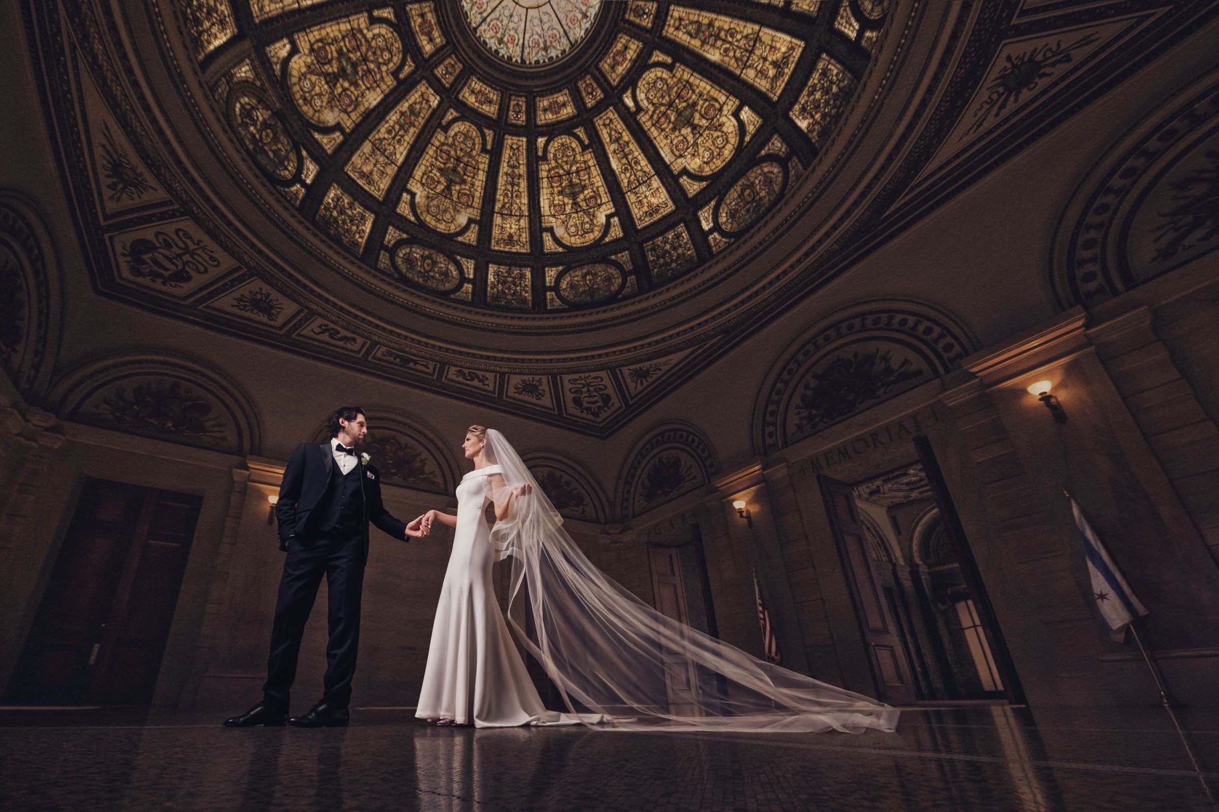 A formal couple stands beneath an ornate domed ceiling with classical architecture and dramatic lighting in a grand ballroom.