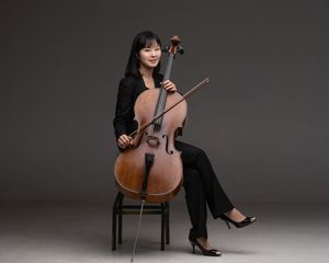 A professional cellist in black attire poses elegantly with their instrument against a dark background for studio portrait.