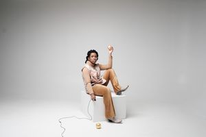 A person in tan clothing sits casually on a chair against a white background, raising one arm in a relaxed pose.