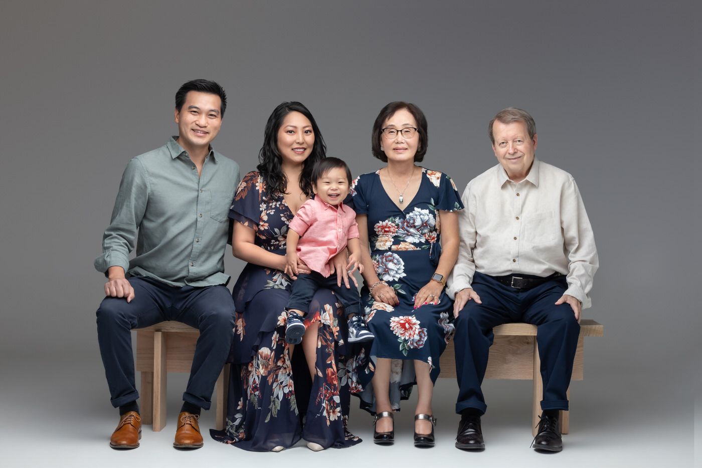 A multi-generational family group poses together for a formal portrait while seated on a bench against a gray backdrop.