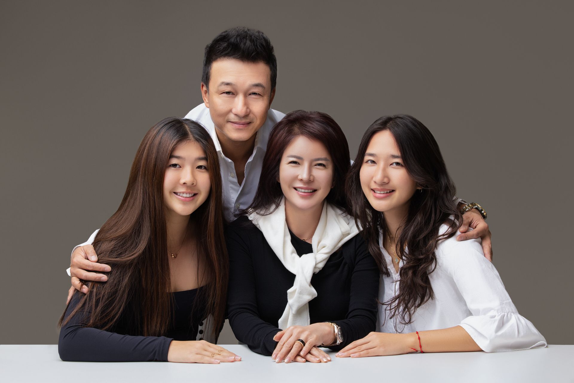 A group of people in white and black clothing lean together and smile warmly against a dark gray background.