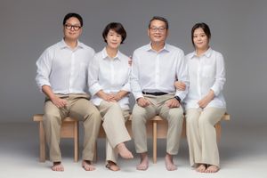 Four people wearing white tops and beige pants sitting together on wooden chairs against a gray background.