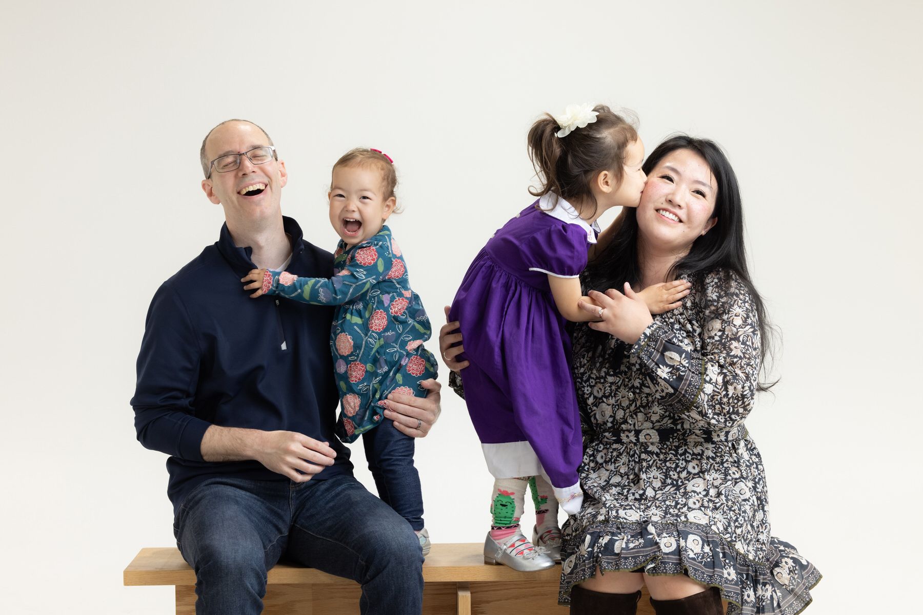 A happy family of four share joyful moments together while posing on a wooden bench against a light backdrop.