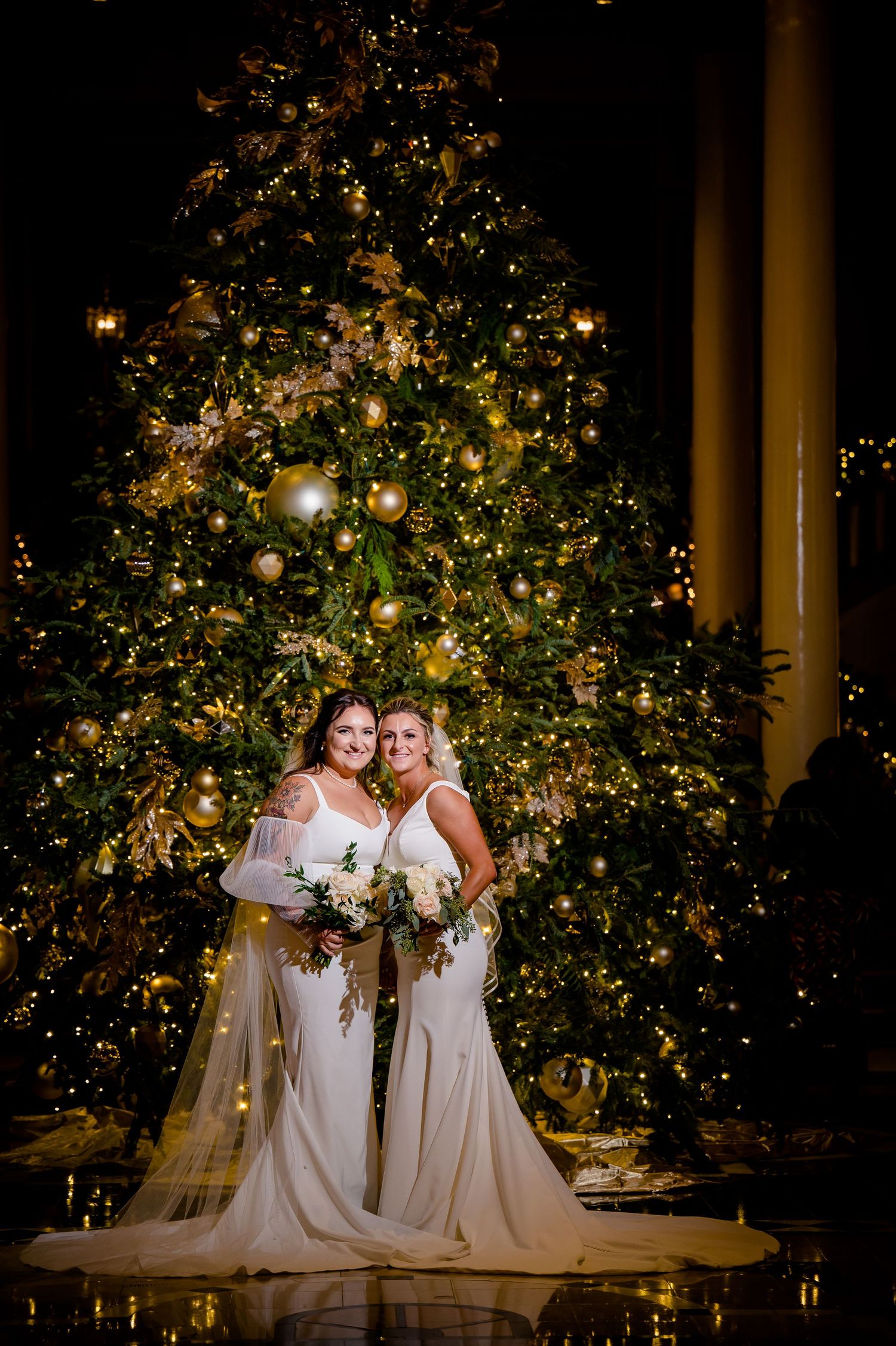 Two brides stand elegantly in front of a beautifully decorated Christmas tree glowing with warm lights and ornaments.