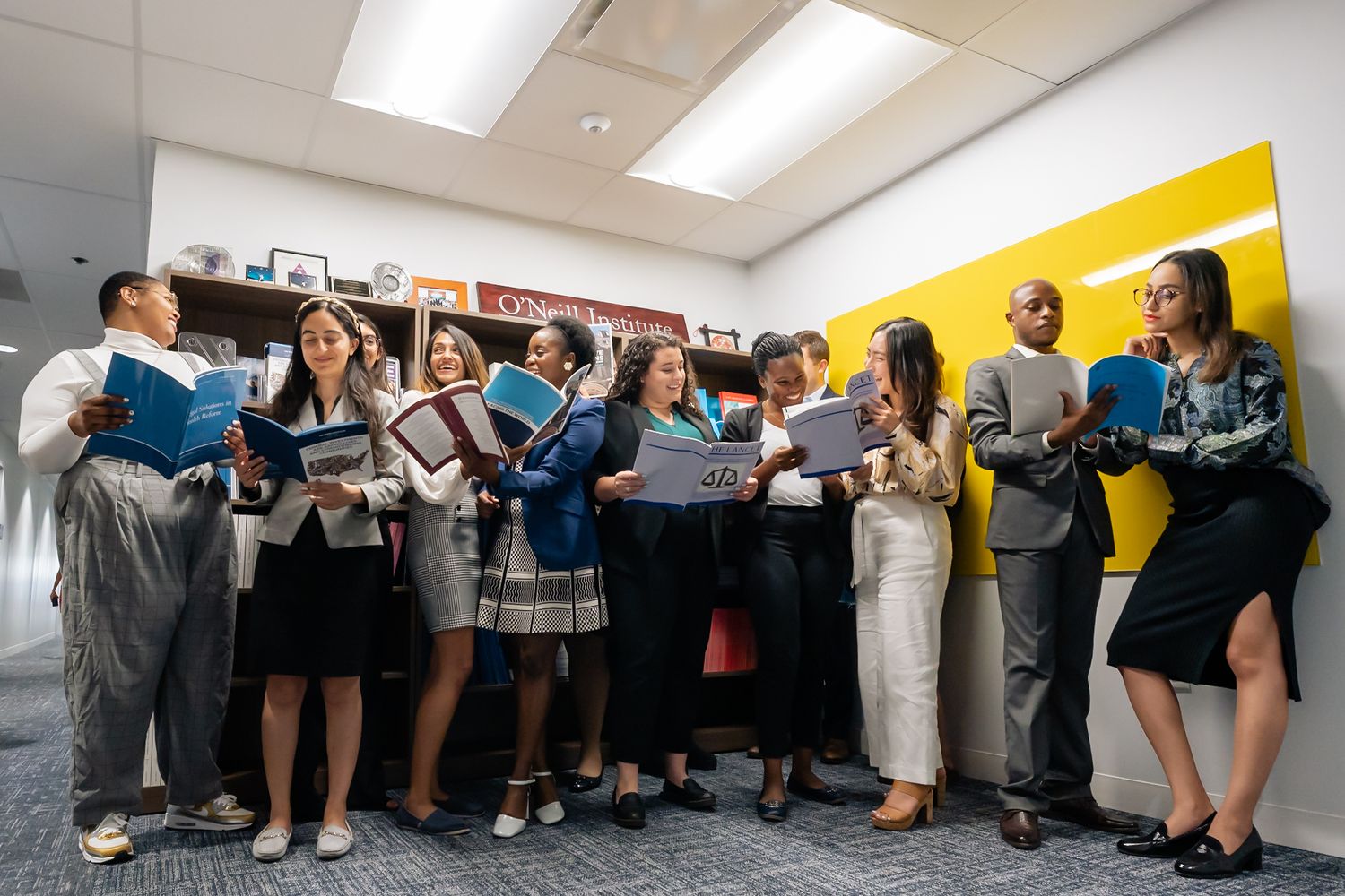 A group of people stand in a line against a wall while reading from blue folders in an office setting.