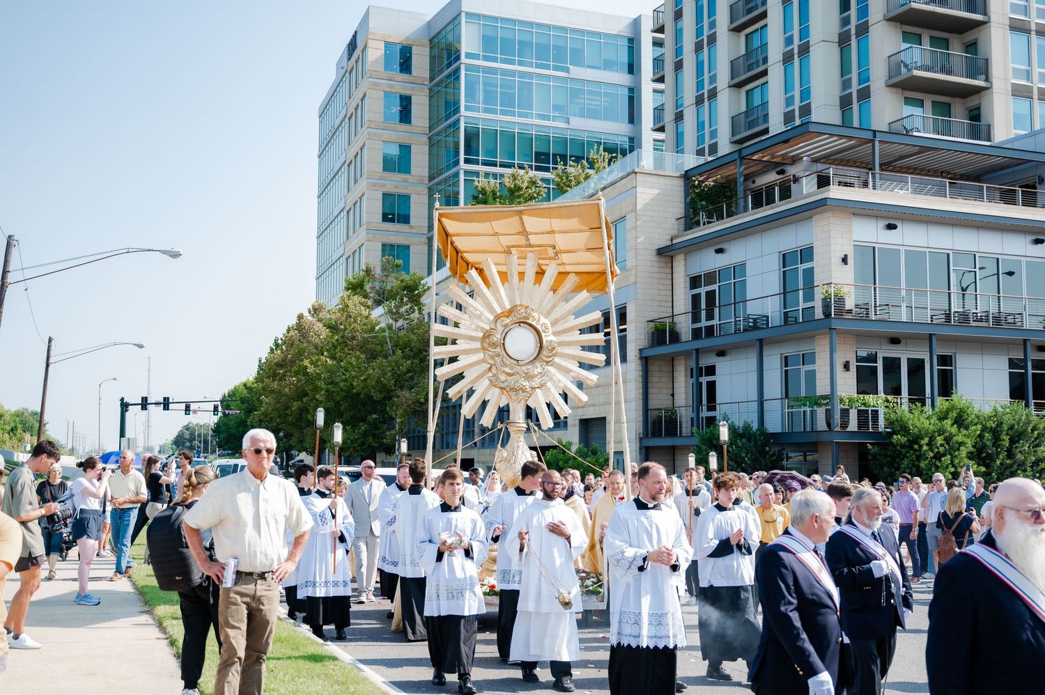 Religious procession through city streets with participants in white robes carrying a golden sunburst banner.