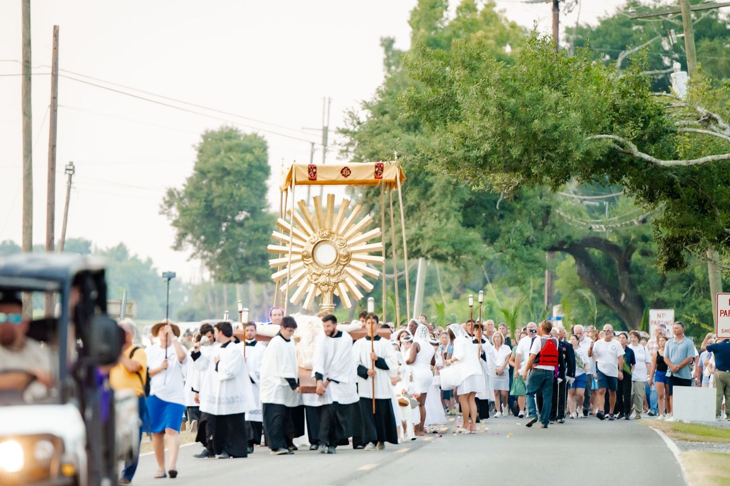 A religious procession with people in white robes carrying a large ornate banner outdoors.
