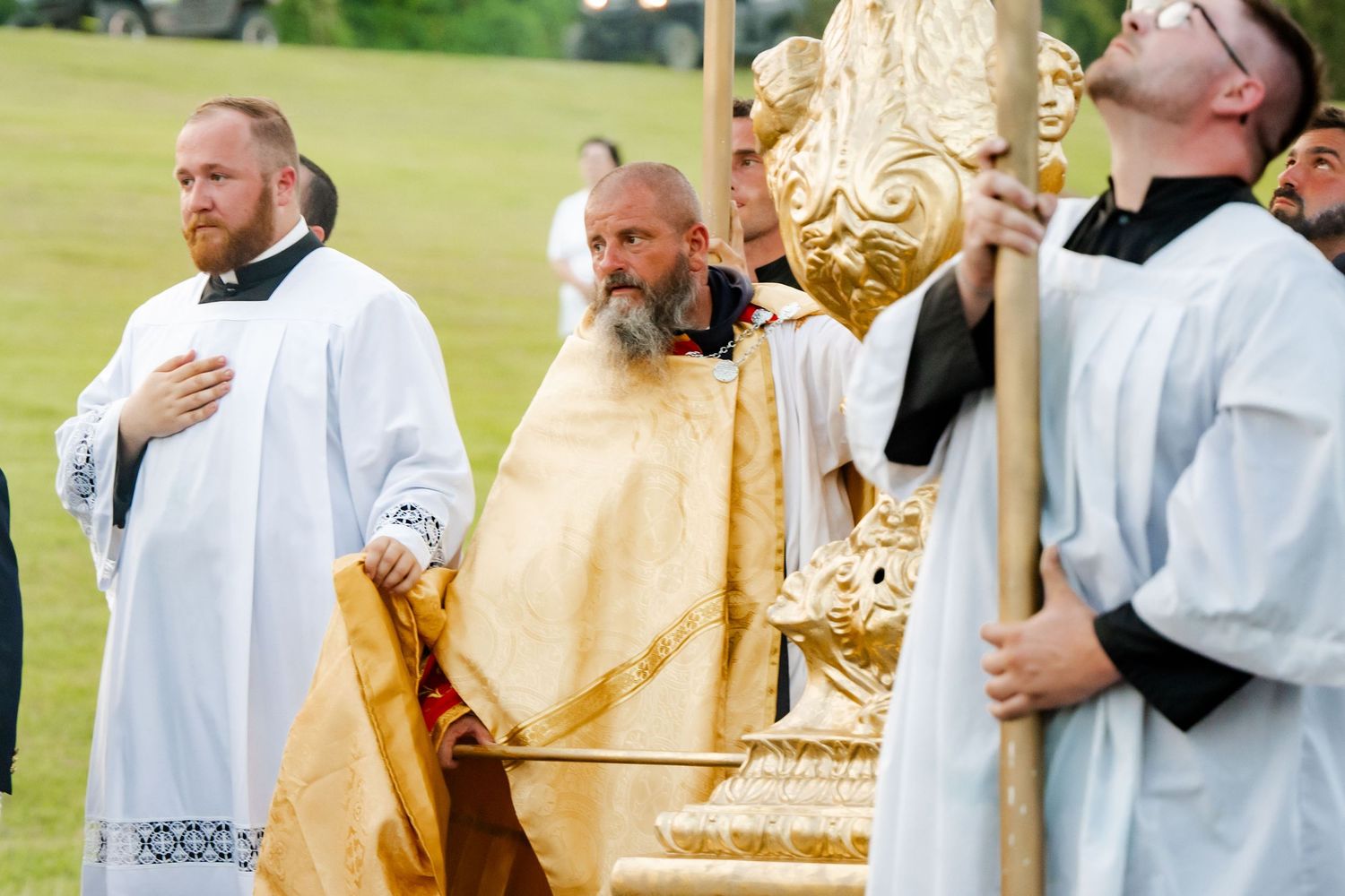 Clergy members in white robes carrying golden religious artifacts during an outdoor ceremony.