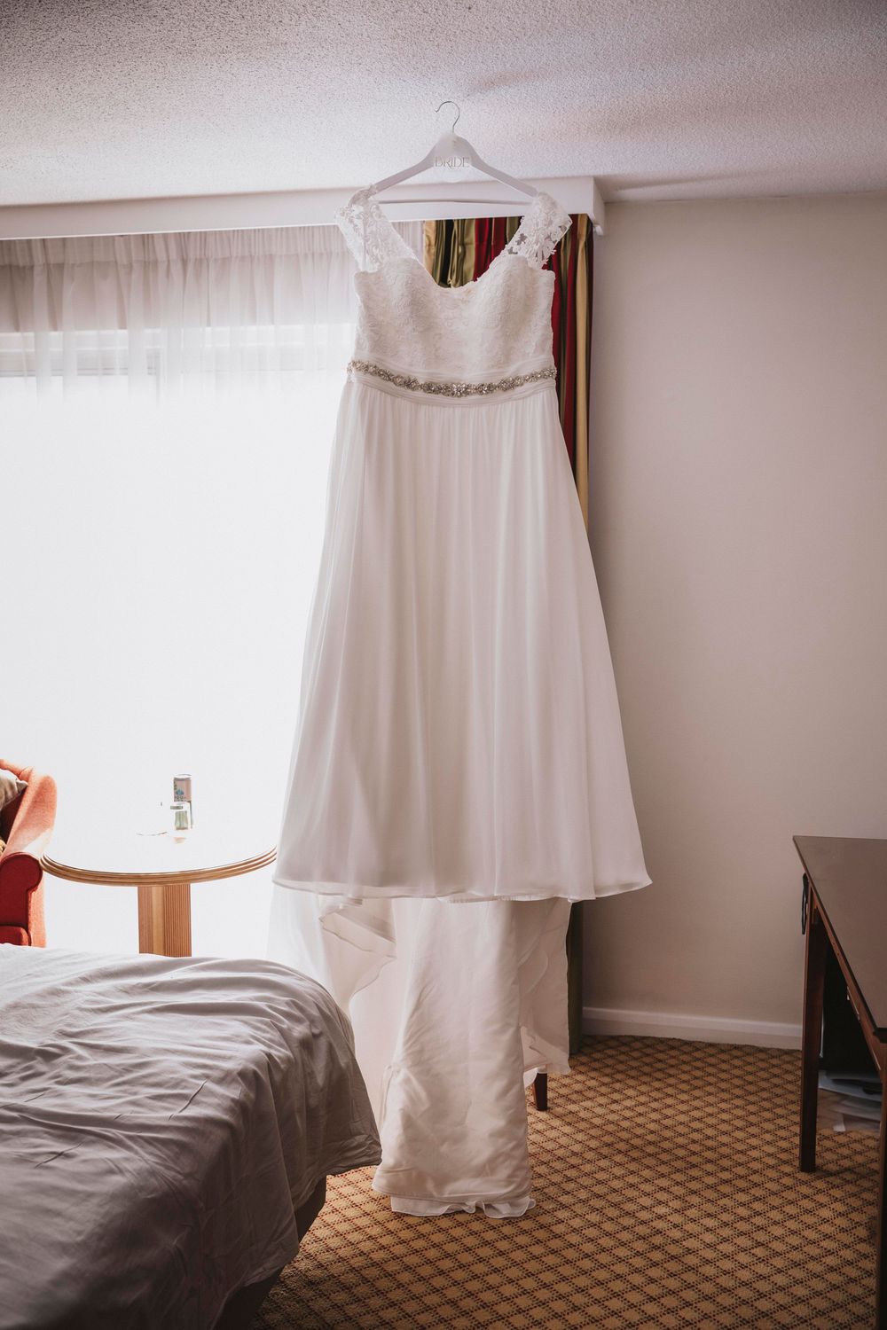 A white wedding dress with delicate details hangs elegantly by a sunlit window in a hotel room.