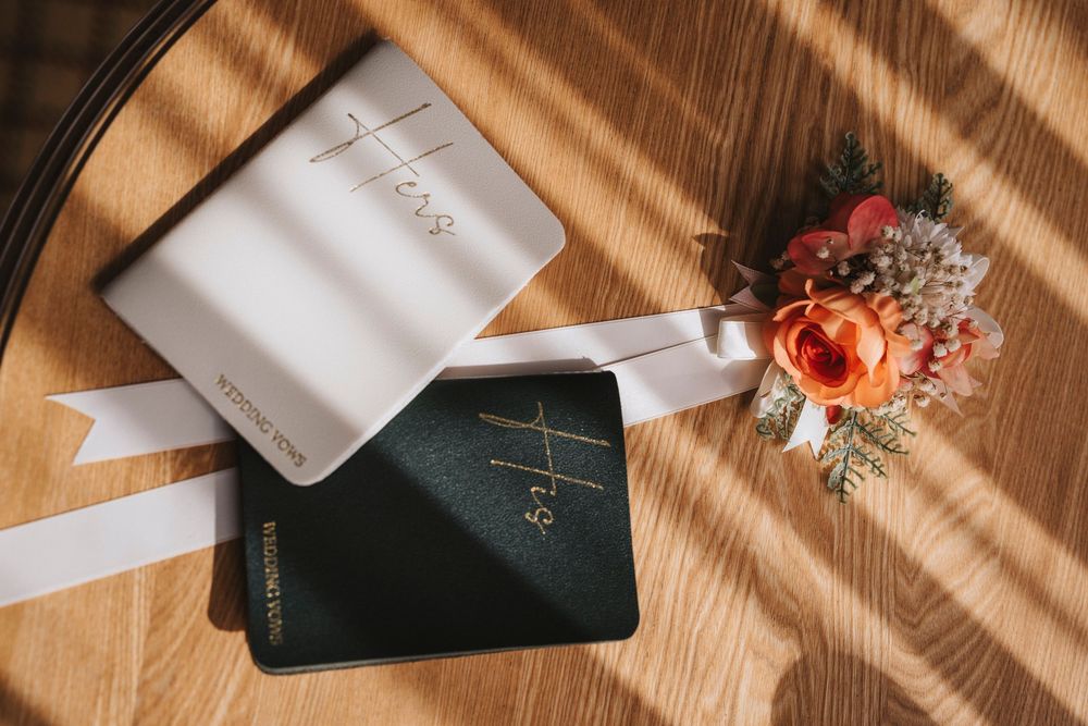 Wedding vows books with 'His' and 'Hers' text lie beside an elegant floral arrangement on a wooden surface.
