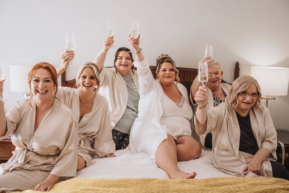 A group of people wearing white robes pose and laugh together during a wedding party preparation.