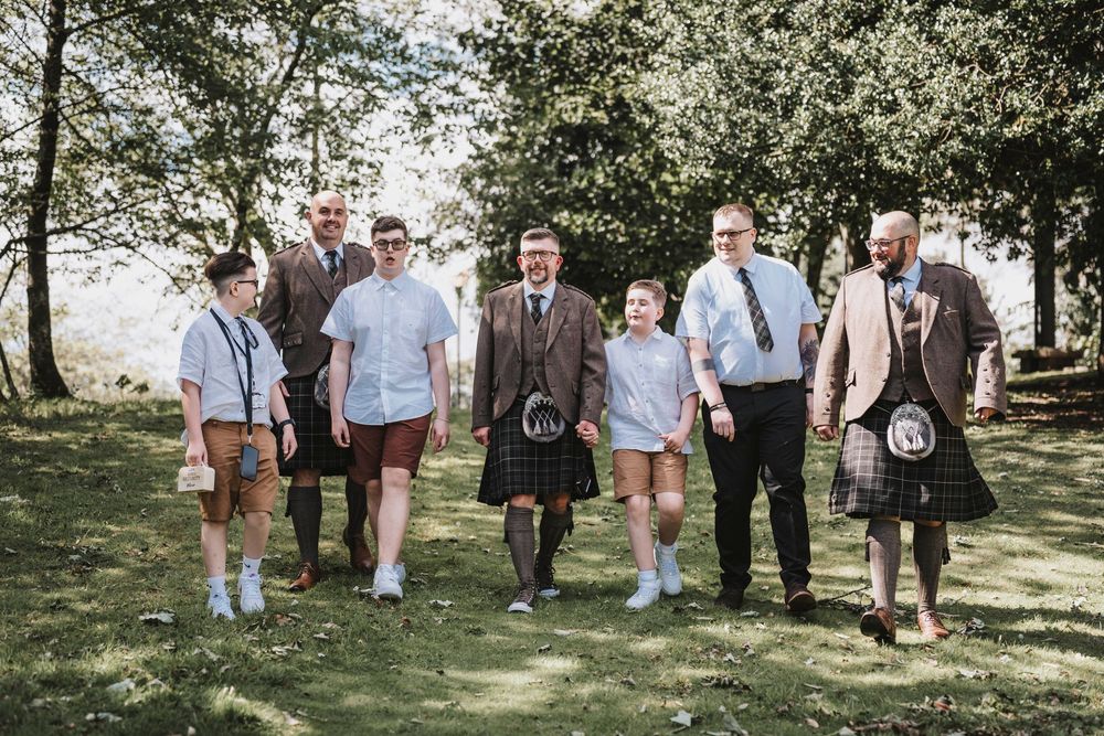 A group of people wearing a mix of traditional kilts and casual clothing stand together outdoors on a sunny day.