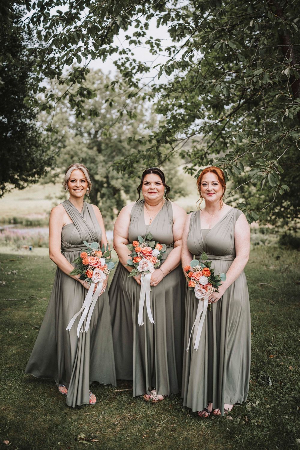 Bridesmaids in sage green dresses holding coral and peach floral bouquets pose together in an outdoor garden setting.