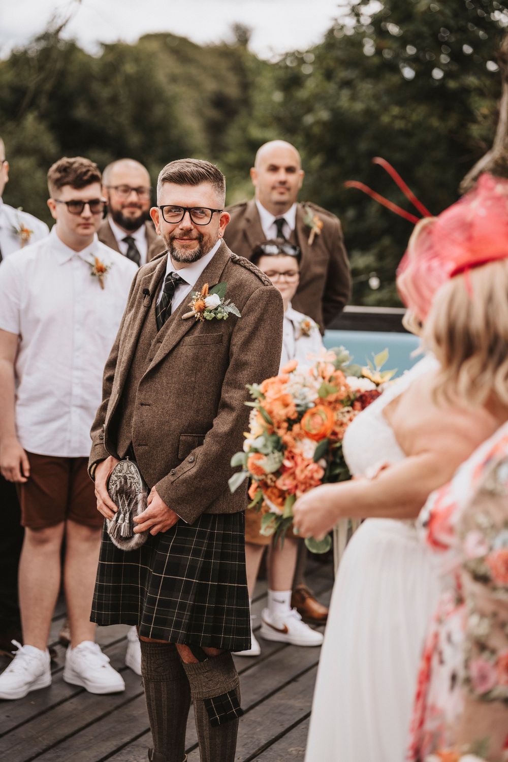 Scottish wedding celebration with groom in traditional tweed jacket and kilt standing on wooden deck with guests.