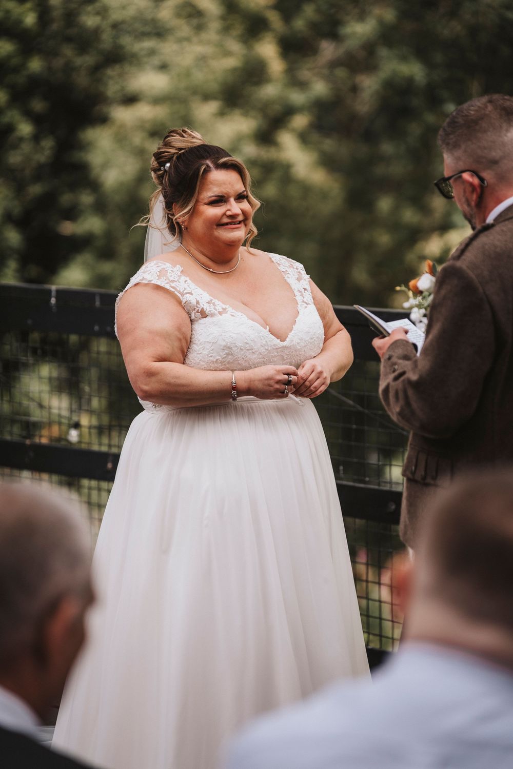 A wedding ceremony takes place outdoors with a couple exchanging vows in front of seated guests during a sunny day.