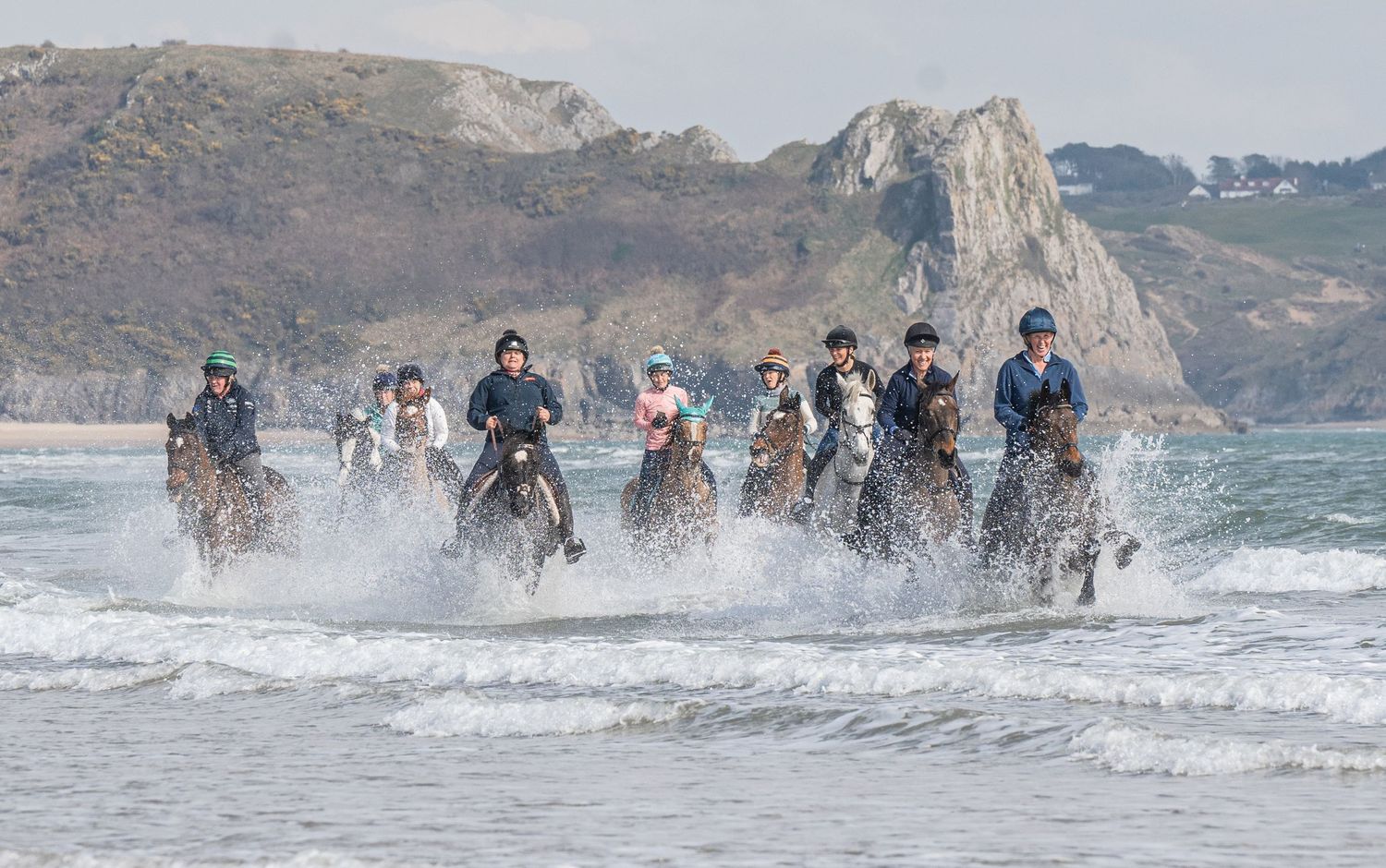 Group of horses and riders galloping through ocean surf with cliffs in background.