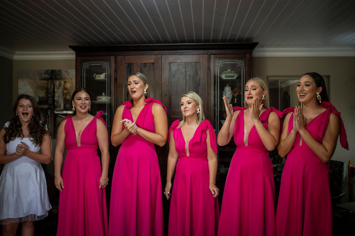 A group of bridesmaids in matching bright pink long dresses react with joyful expressions at a bridal event.
