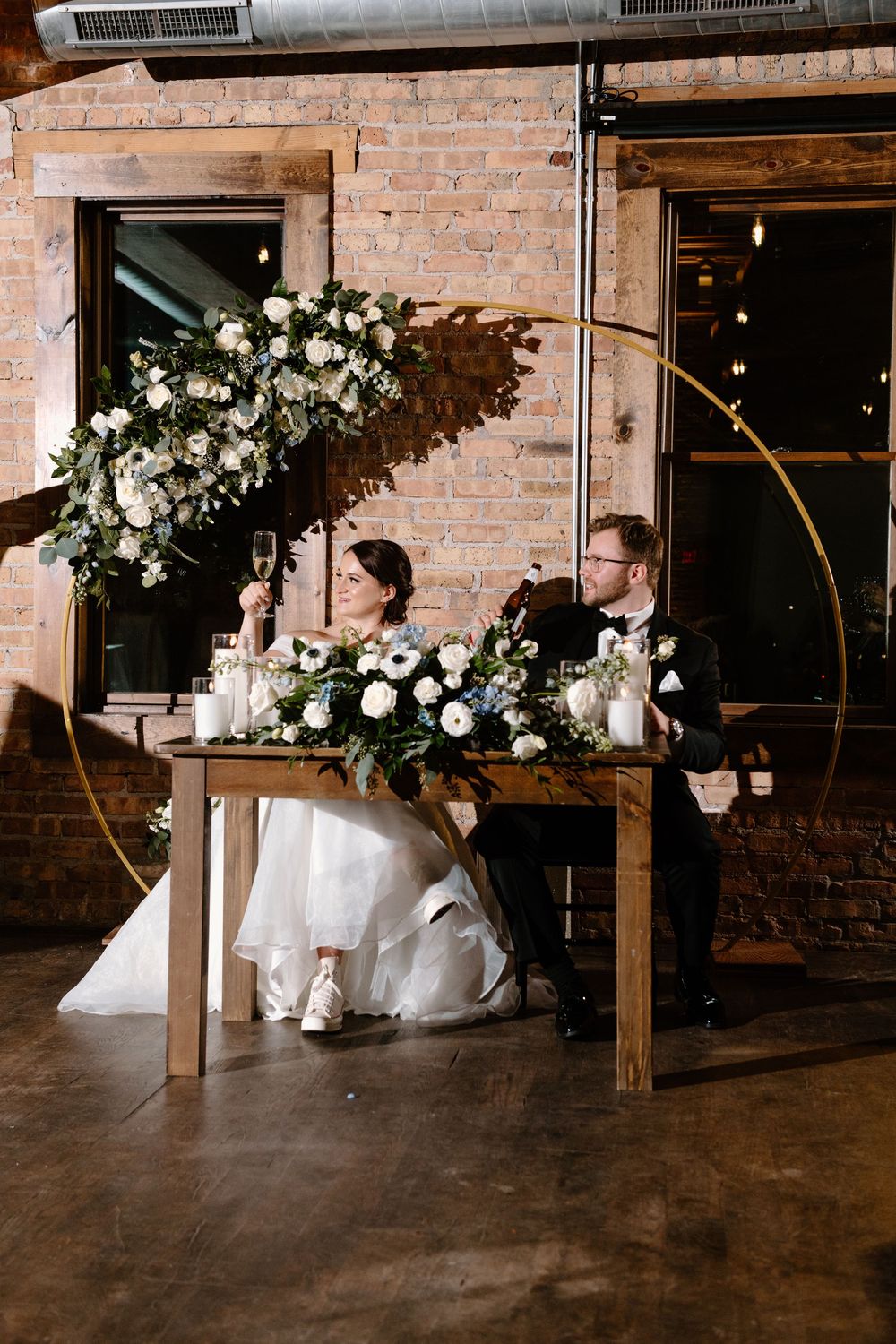 A newlywed couple sits at a wedding reception table decorated with white floral arrangements and a gold arch detail.