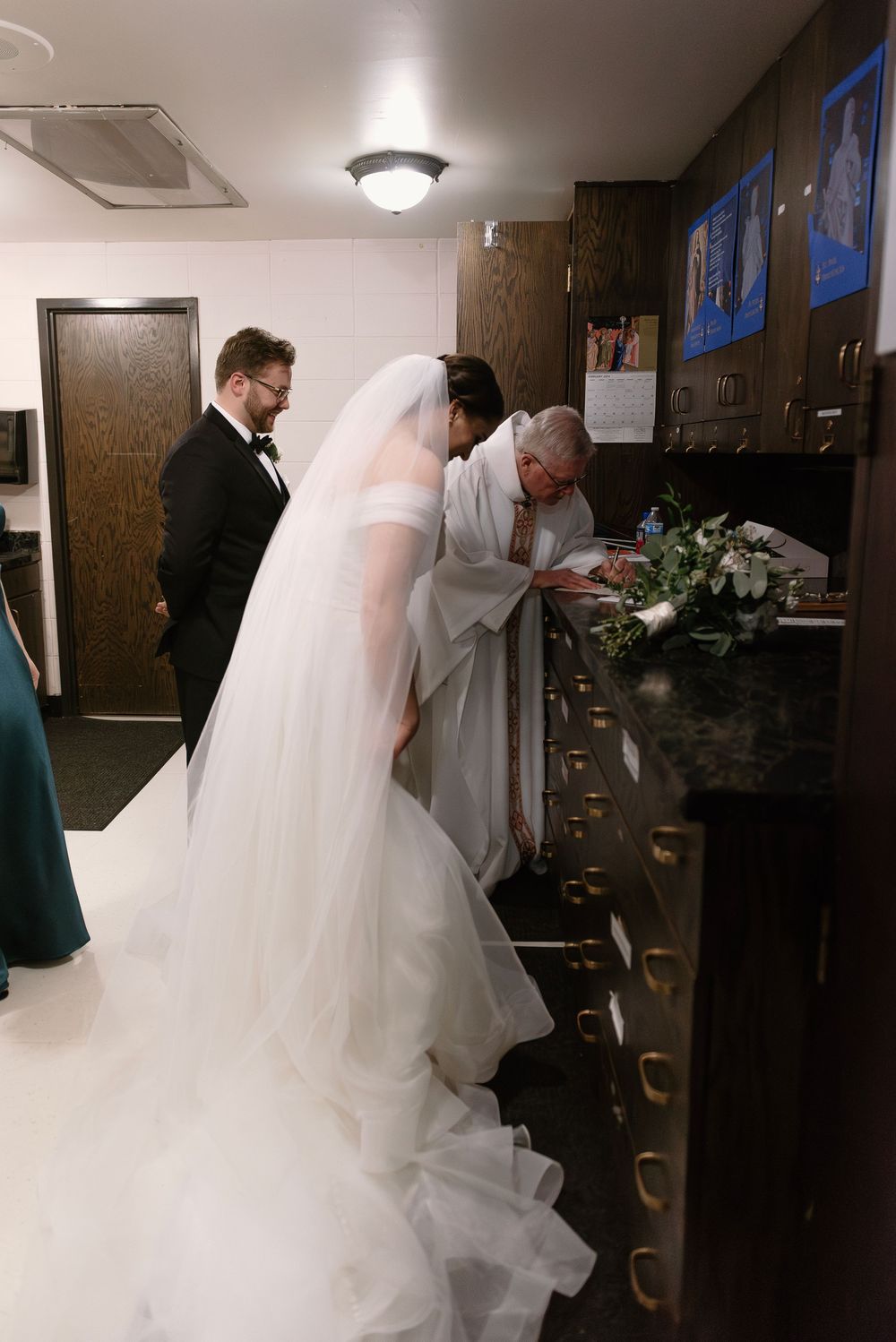 Bride in white gown and veil signing wedding documents with groom and officiant at a church registry.