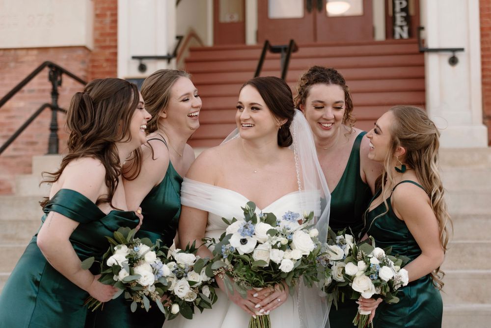 Bride in white gown laughing with four bridesmaids in emerald green dresses holding floral bouquets.