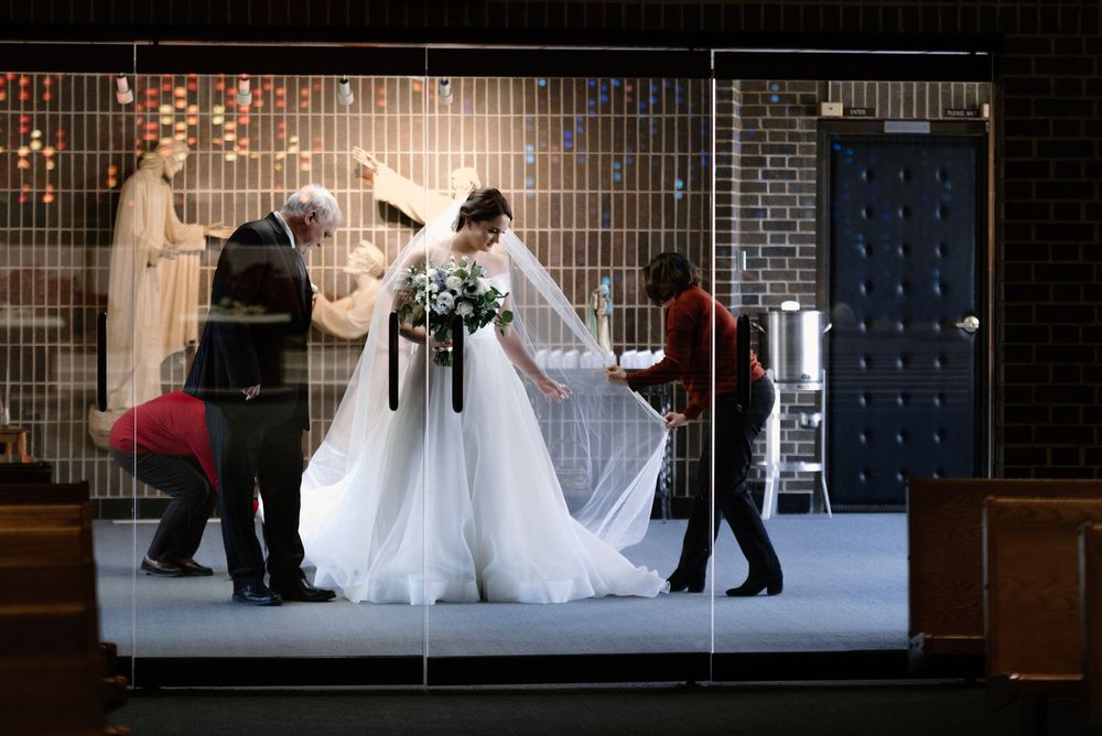 Bride in white gown with veil being attended by helpers inside a church before wedding ceremony.