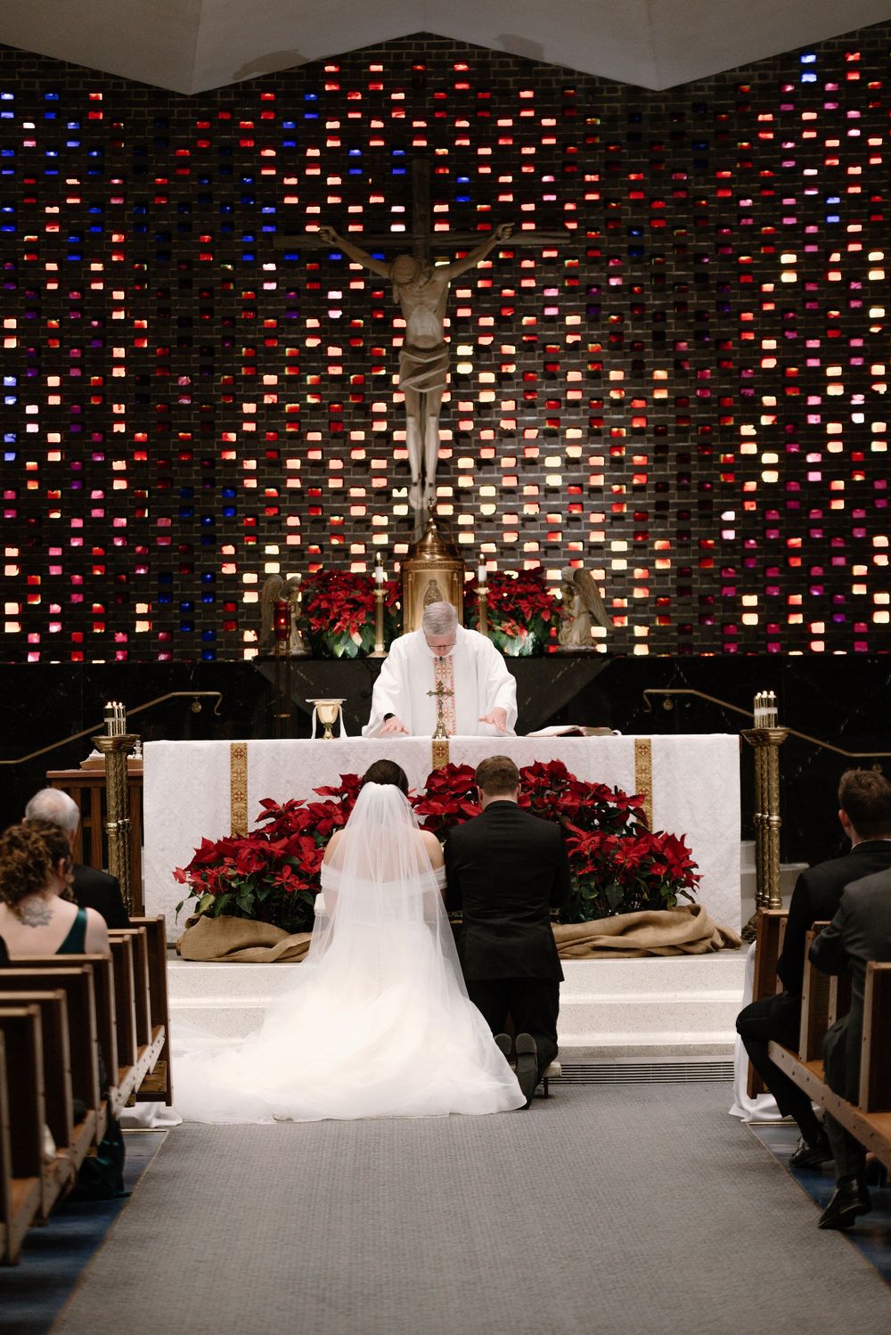 Bride and groom kneeling at church altar during wedding ceremony with priest and red poinsettias.