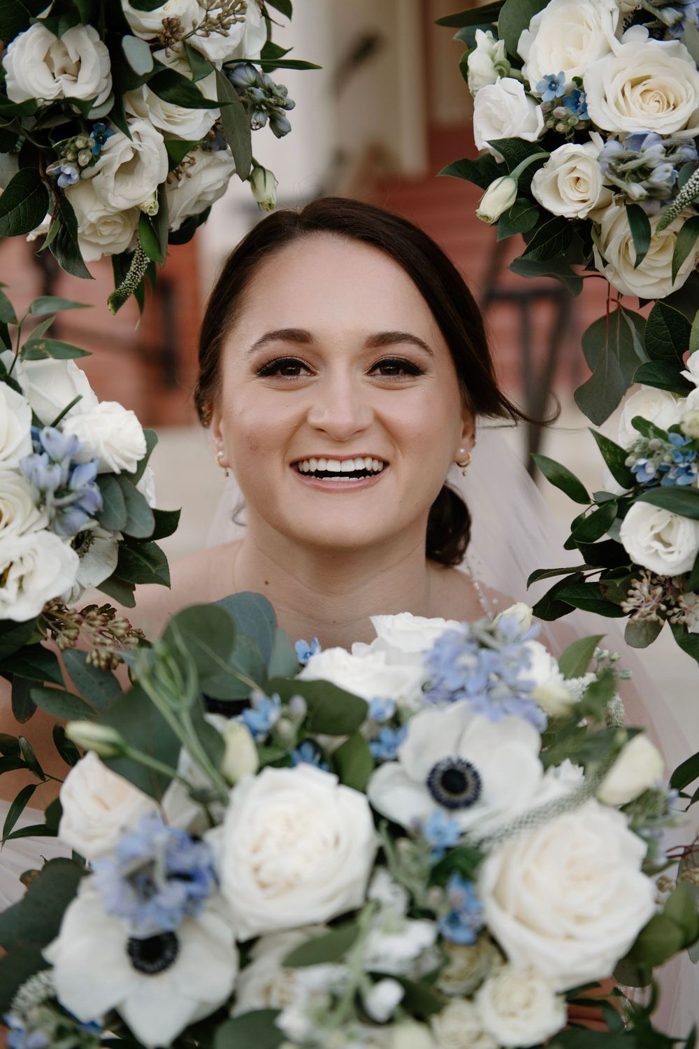 Smiling bride framed by white and blue floral arch, holding bouquet of roses and anemones on her wedding day.