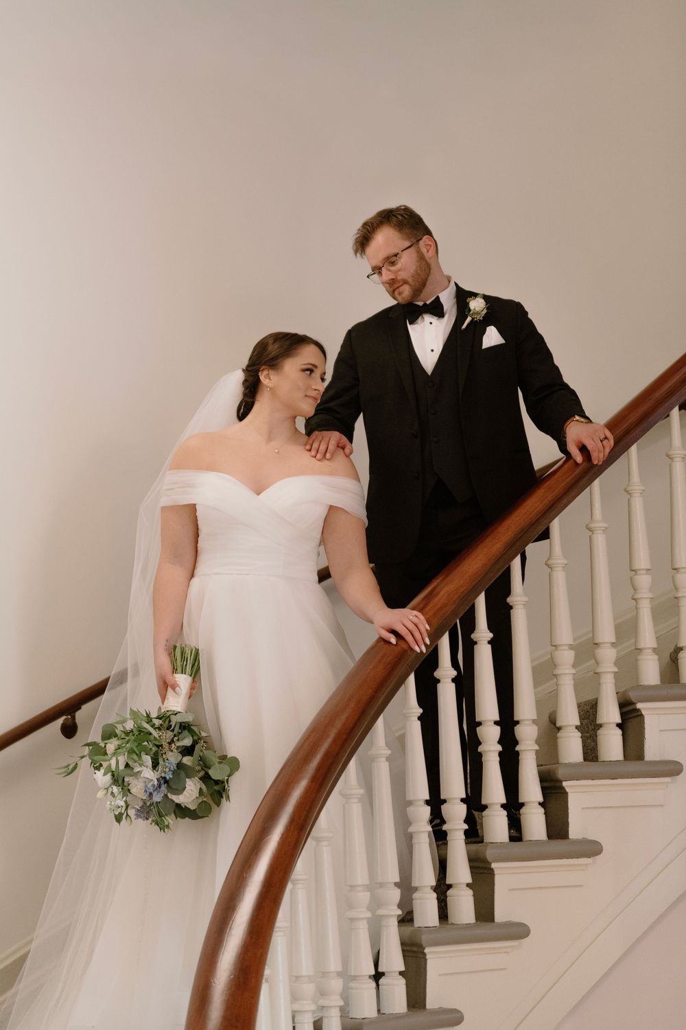 Bride in off-shoulder gown with bouquet and groom in black tuxedo pose on elegant staircase.