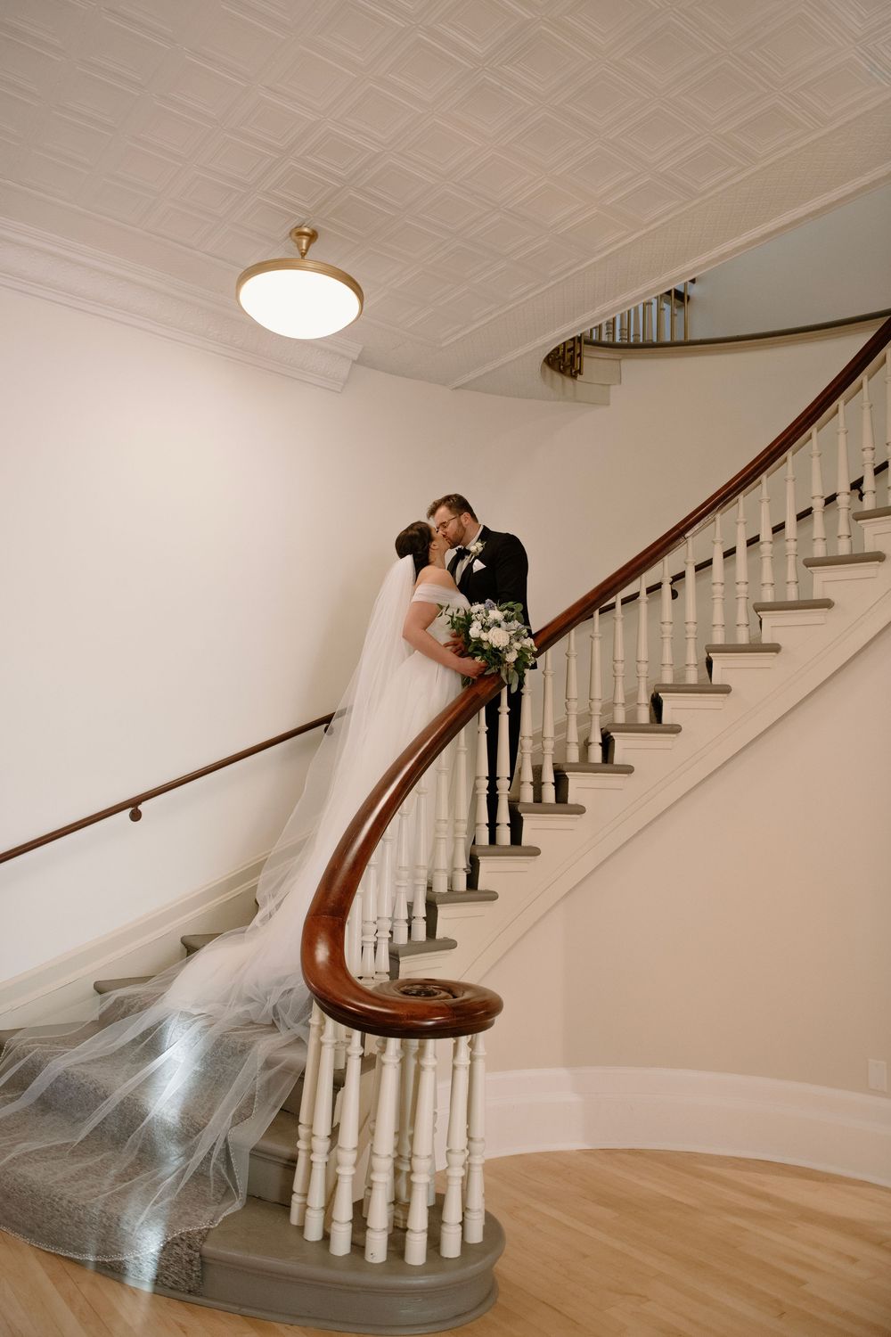 Bride and groom share a romantic kiss on an elegant curved staircase with white balusters and mahogany railing.