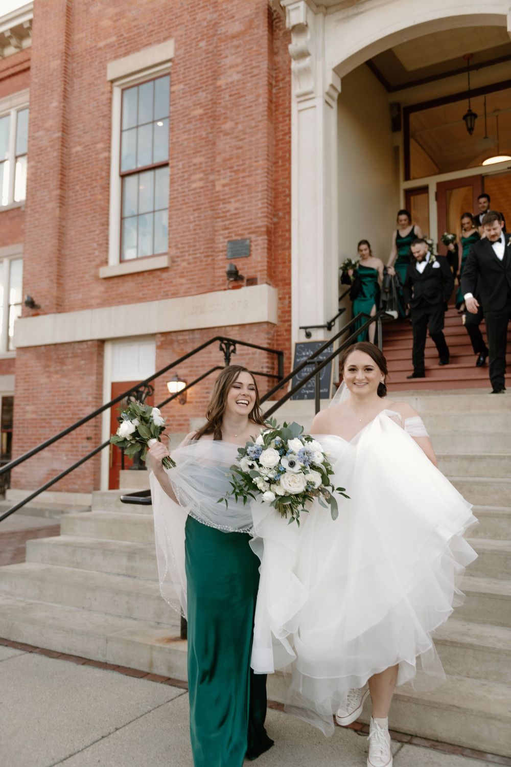 Bride in white gown with bouquet walks down brick building steps with bridesmaid in green dress at wedding.
