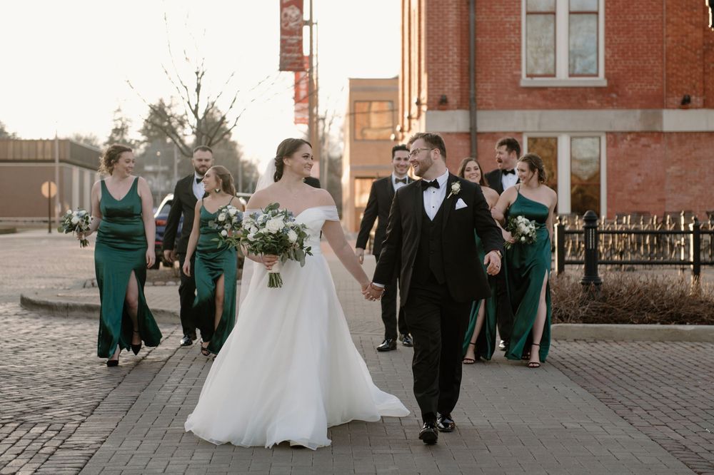 Bride and groom walk with wedding party in emerald green dresses and black tuxedos on a brick street.
