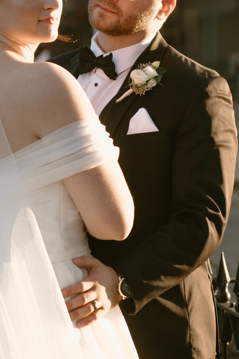 Bride in off-shoulder gown and groom in black tuxedo with white boutonniere embrace at golden hour wedding.