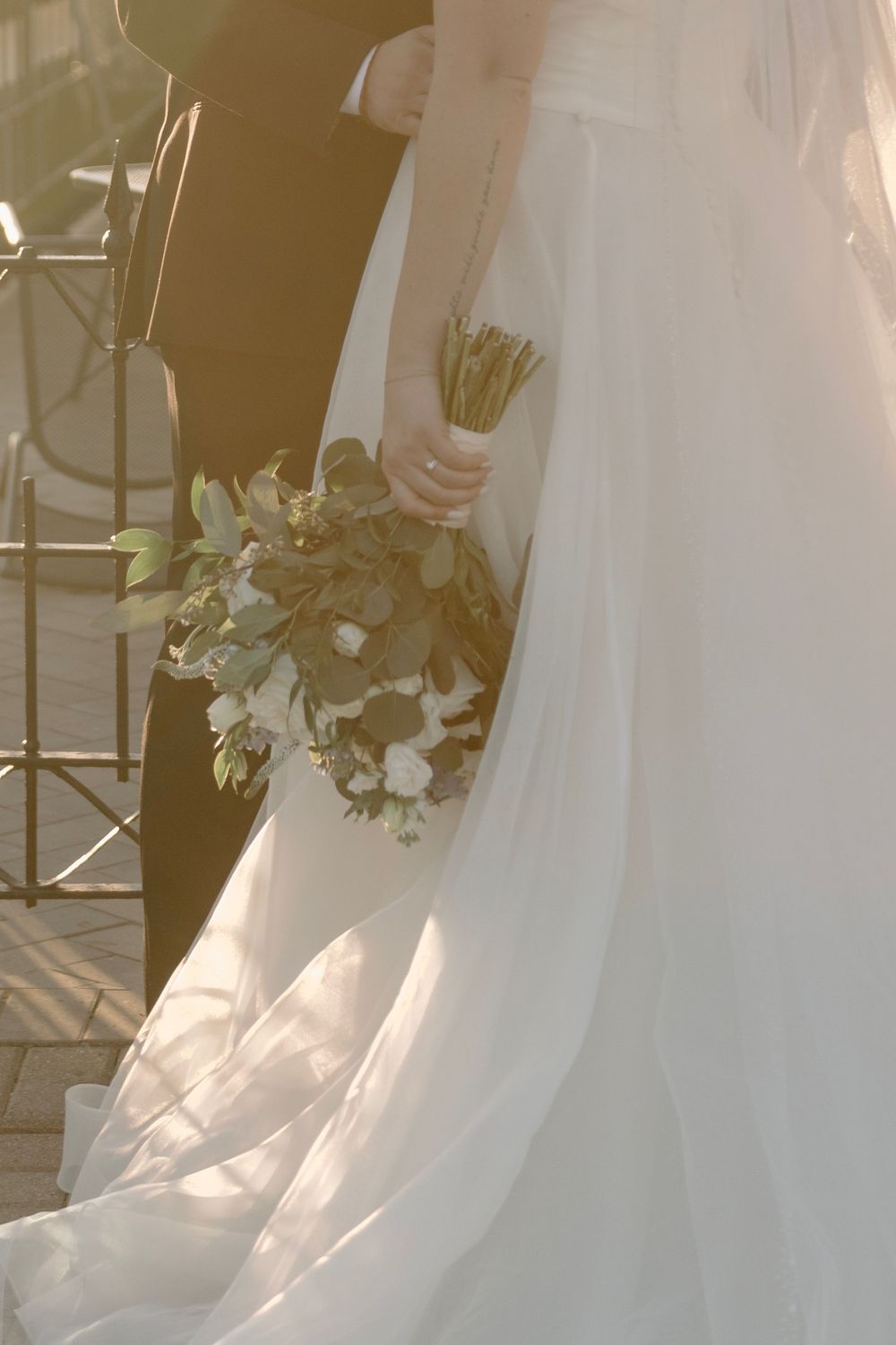 Bride in flowing white gown holding a green and white bouquet, bathed in warm golden sunlight.