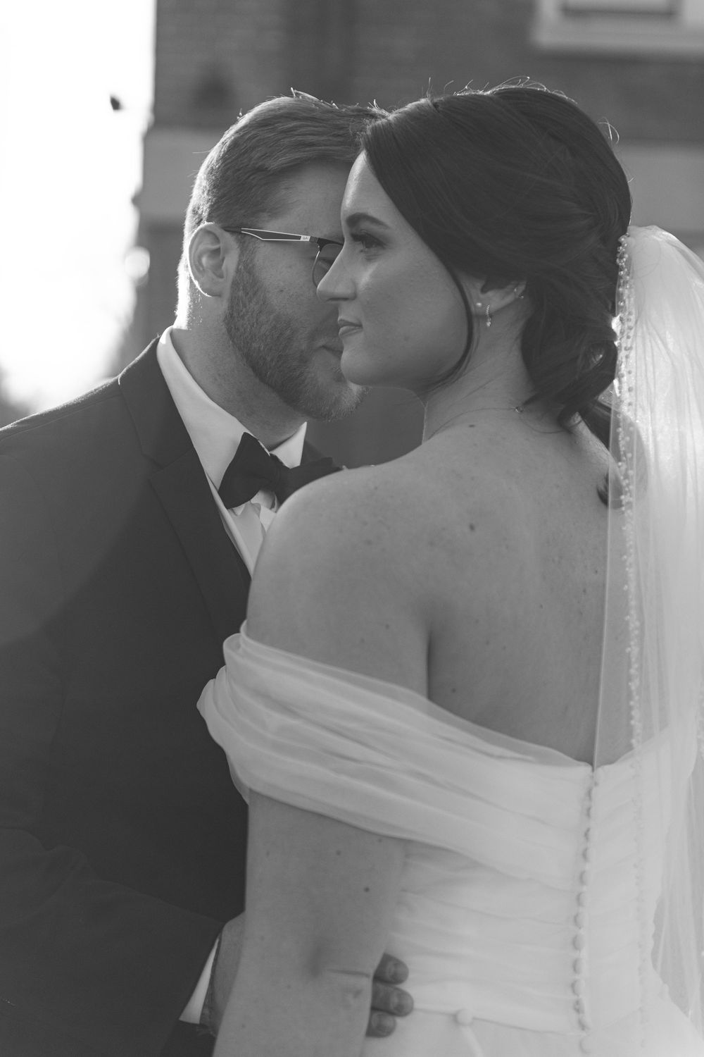 Black and white wedding photo of bride in off-shoulder gown with veil and groom in tuxedo sharing an intimate moment.