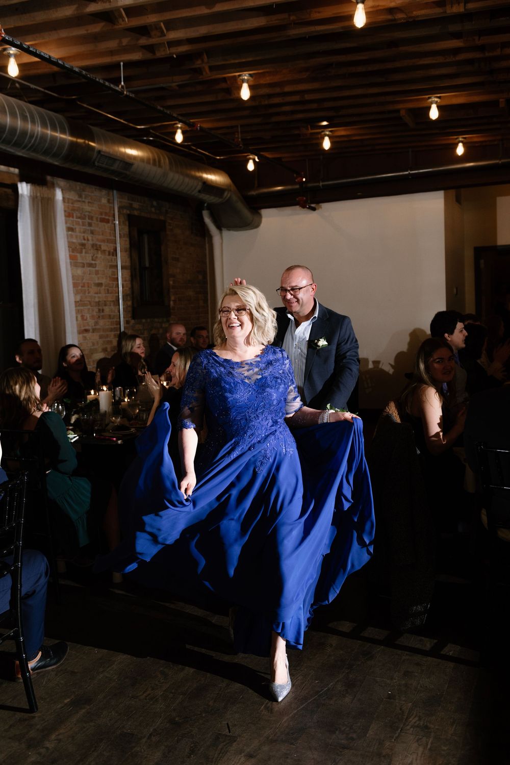 Woman in royal blue gown twirls on dance floor at elegant wedding reception with string lights.
