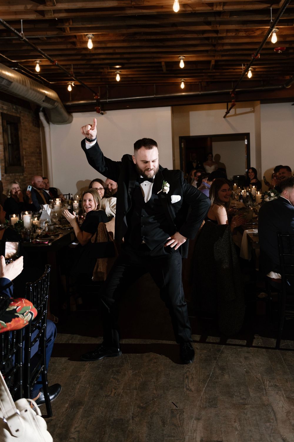 Groom dancing energetically at wedding reception in black tuxedo, guests cheering in warmly lit venue.