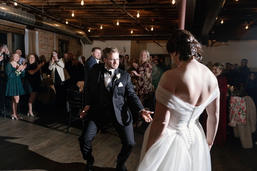 Groom excitedly greets bride during wedding reception grand entrance in rustic venue with exposed brick and Edison bulb lighting.