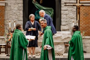 Clergy members in green robes interact with attendees during a church ceremony near stone walls and brick architecture.