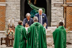 Group of clergy members wearing bright green robes walk in a line as others wave in the background by stone walls.