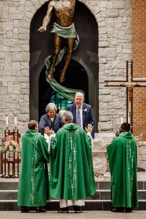 Religious clergy members in green robes stand outside a stone church with their hands raised during a ceremony.
