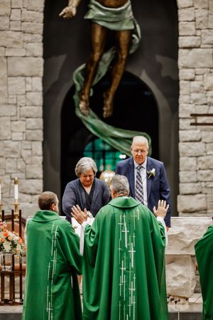 Clergy members in green robes perform a blessing ceremony inside a stone church with dramatic religious artwork above.