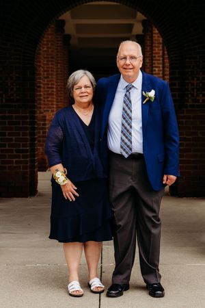 An elderly couple in formal attire stand together in front of a brick building entrance.