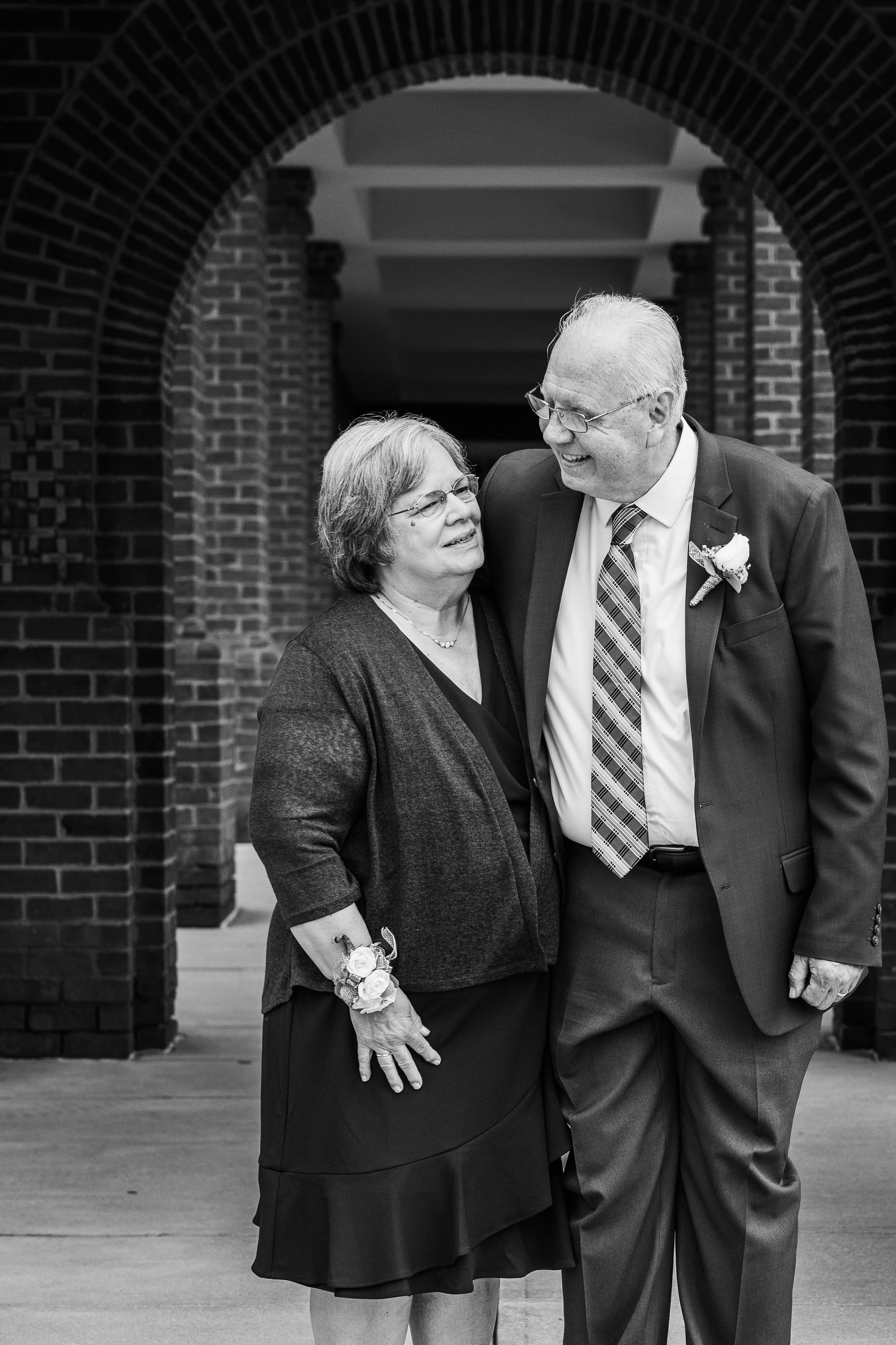 A couple dressed in formal attire shares a tender moment while standing against a brick archway background.
