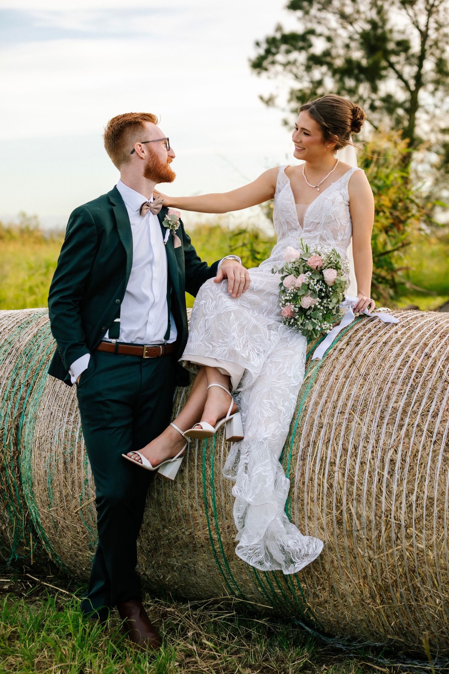 A couple in wedding attire poses together on a hay bale in a rustic outdoor setting during golden hour. Queensland Wedding Photography
