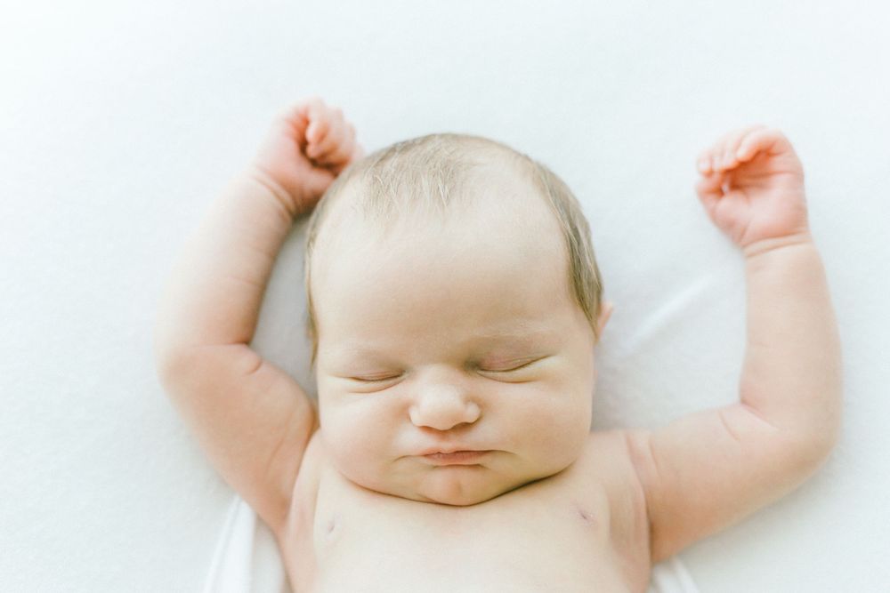 Baby peacefully sleeping with arms outstretched in white wrap on light background.