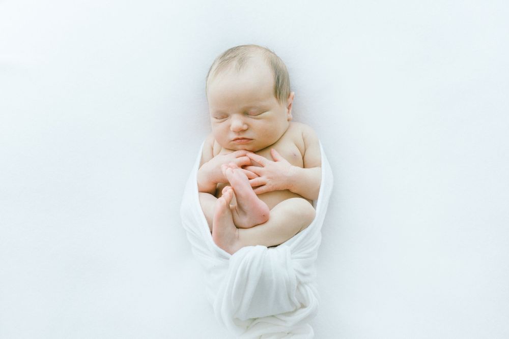 Newborn swaddled in white fabric resting against clean white backdrop.