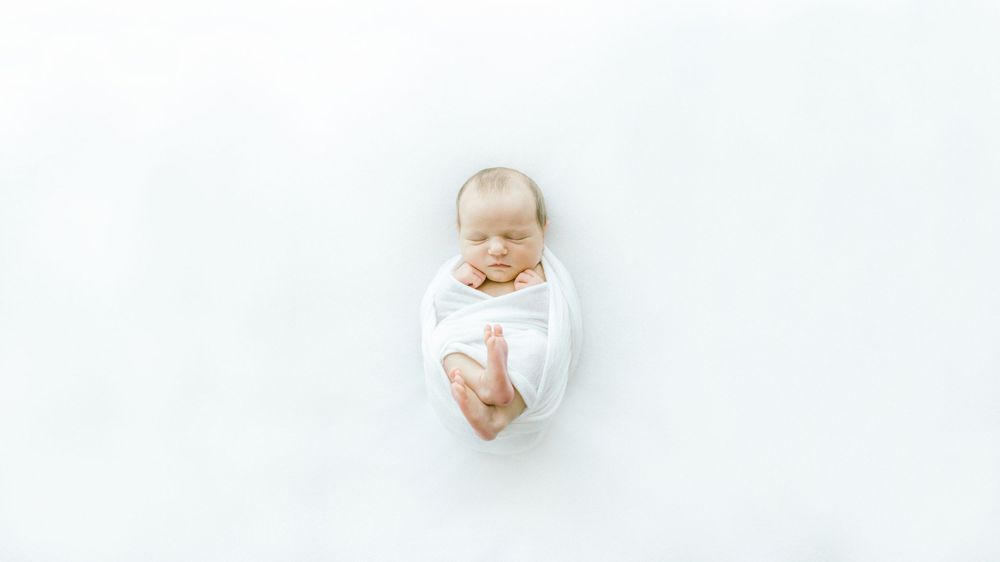 Infant wrapped in white blanket with gentle expression on white backdrop.