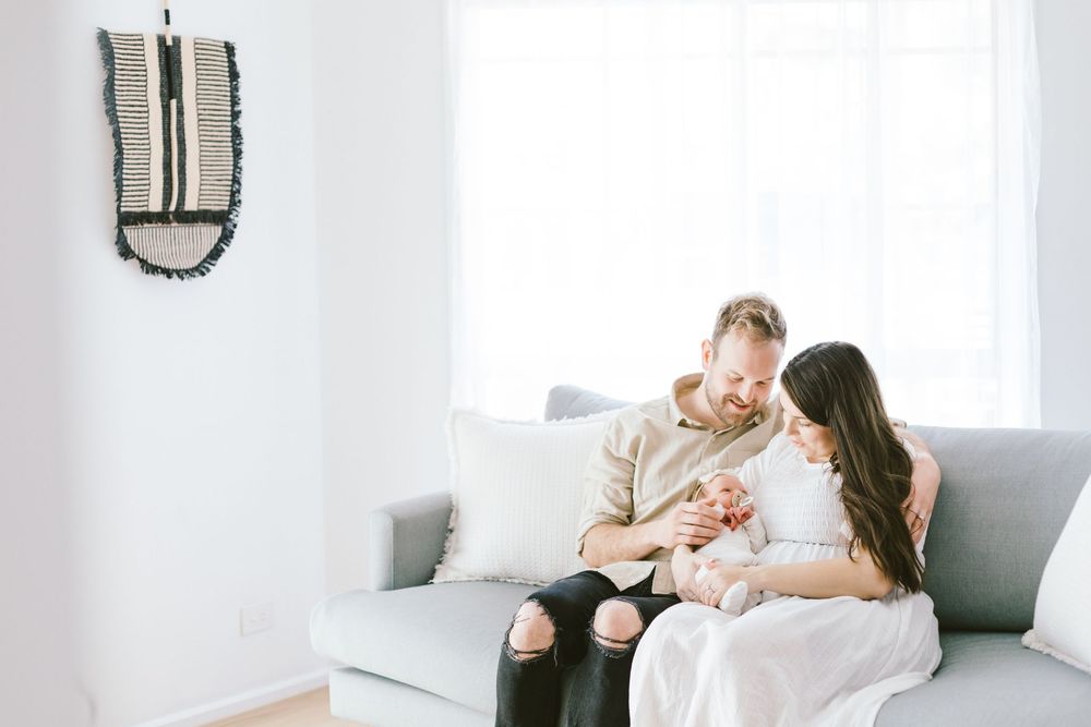 Parents and chocolate labrador dog share tender moments with their newborn baby on white couch.
