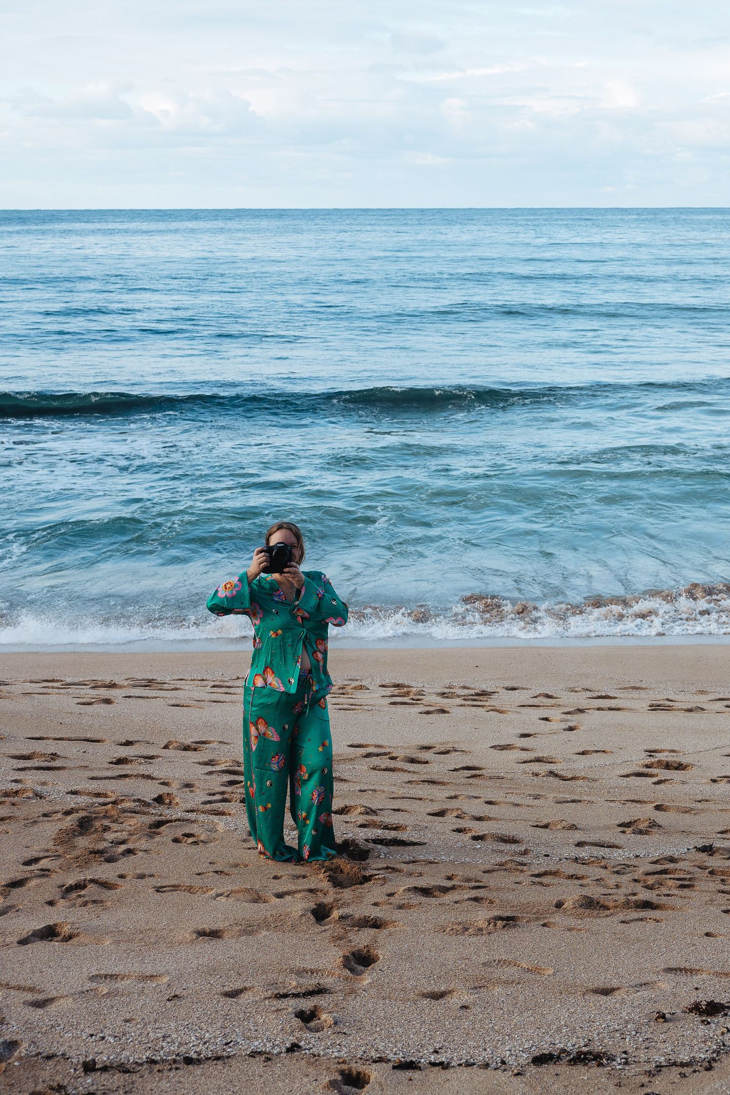 Person in green floral outfit stands on sandy beach taking photo of blue ocean waves and horizon.